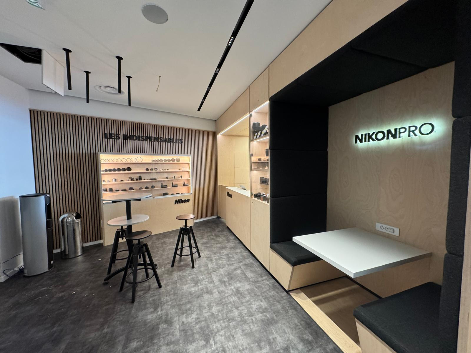 Intérieur de la boutique Nikon Pro : enseigne NIKONPRO rétroéclairée, mur d'exposition en tasseaux de bois et coin salon intime.